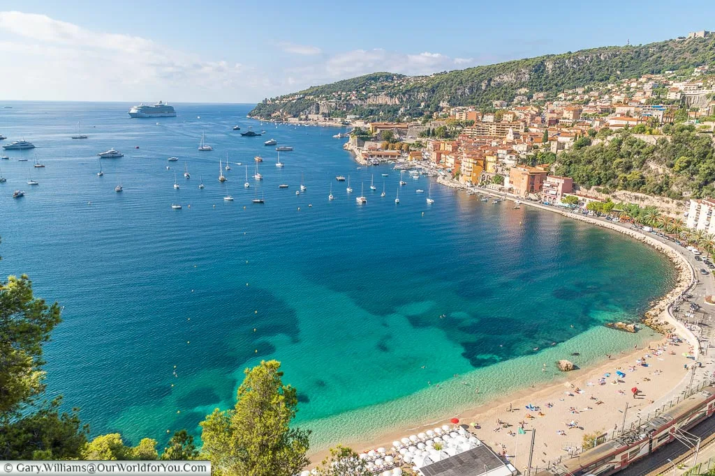 The topaz blue waters of the bay of Villefranche-sur-Mer as seen from above with the beach in the foreground. To the right is the town itself, and Villefranche-sur-Mer is full of small sailing boats.