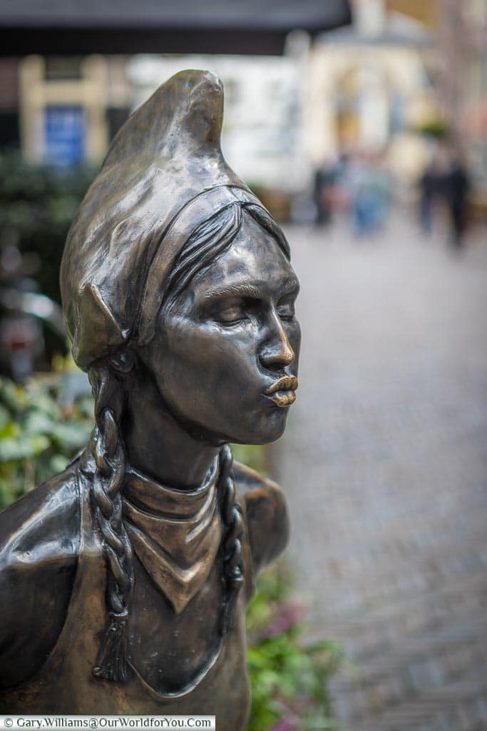 A brass statue of a young Dutch girl in traditional dress with her eye closed, lips puckered ready for a kiss in the centre of Alkmaar in the Netherlands