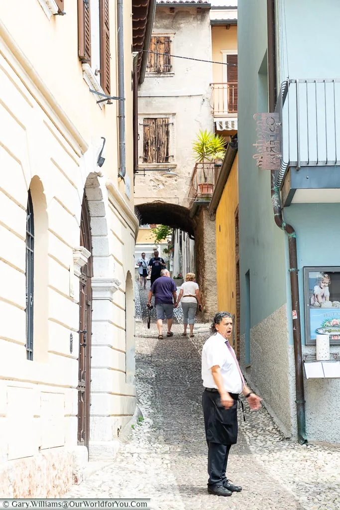 A narrow lane is heading away from the lakeside uphill. At the base of the hill is a waiter trying to drum up trade for his restaurant.