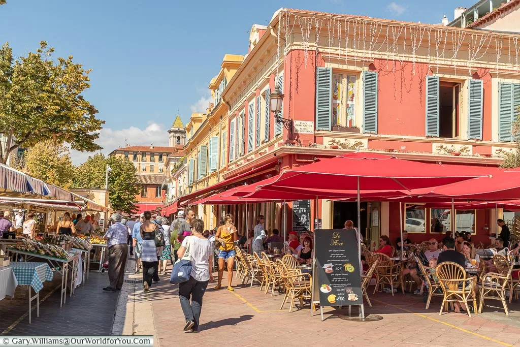 A street scene of a café next to the fruit & vegetable market with people sitting at a bunch of tables under bright red canopies.