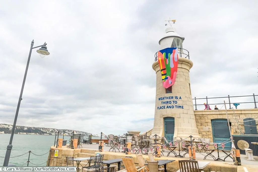 Folkestone Lighthouse, now a bar with tables & chairs outside decorated with bunting. The lighthouse is also adorned with the Creative Artworks Folkestone flag and the inscription "Weather is a third to Place and Time."