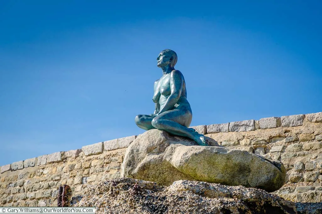 Looking up at the life-sized bronze figure of the Folkestone Mermaid perched on rocks at the edge of Folkestone's Sunny Sands beach
