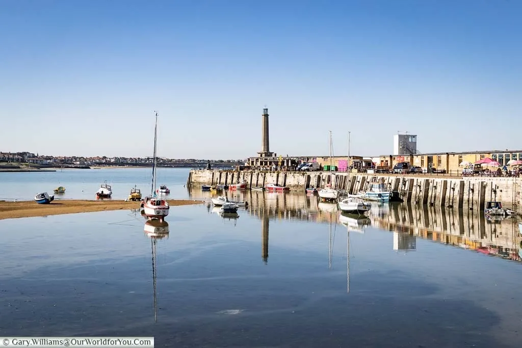 The Harbour at Margate on a bright day at low tide. Some boats are stranded on the sand banks and others partially floated. You can see the Harbour arm leading to the lighthouse at the tip of the Harbour’s edge.