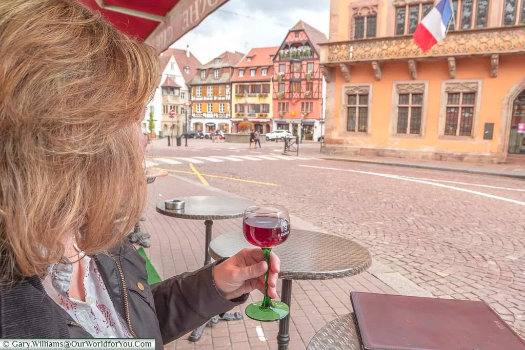 Janis holding a glass of pinot noir wine in Obernai, France. As is tradition in the region the wine glass has a green glass stem and a clear top. She is overlooking the town square including the town hall displaying the French tricolore.