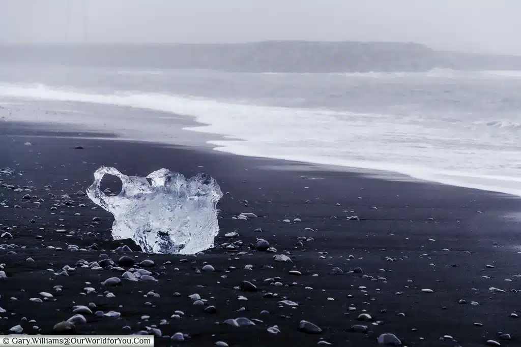 A lump of crystal clear ice washed up on the jet black sand of Diamond Beach with the rolling waves in the background.