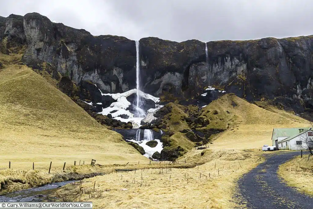 A pair of narrow waterfalls making a significant drop to the river below, next to an Icelandic farmhouse