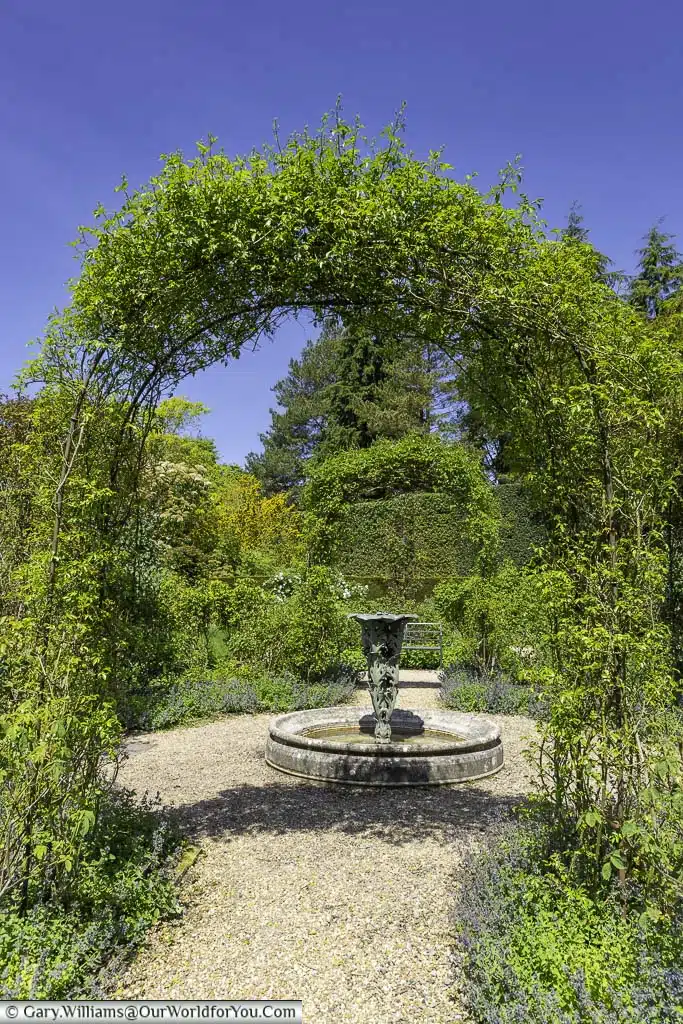 A gravel footpath running under a rose arch towards an art deco water fountain in nymans rose garden