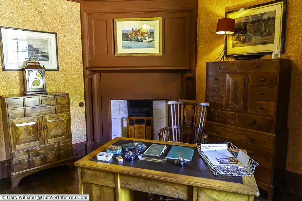 Patterned wallpaper walls on either side of the fireplace set in a rust red wall behind the desk in the business room at standen house in west sussex