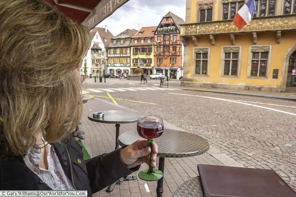 Janis holding a glass of pinot noir wine in Obernai, France. As is tradition in the region the wine glass has a green glass stem and a clear top. She is overlooking the town square including the town hall displaying the French tricolore.