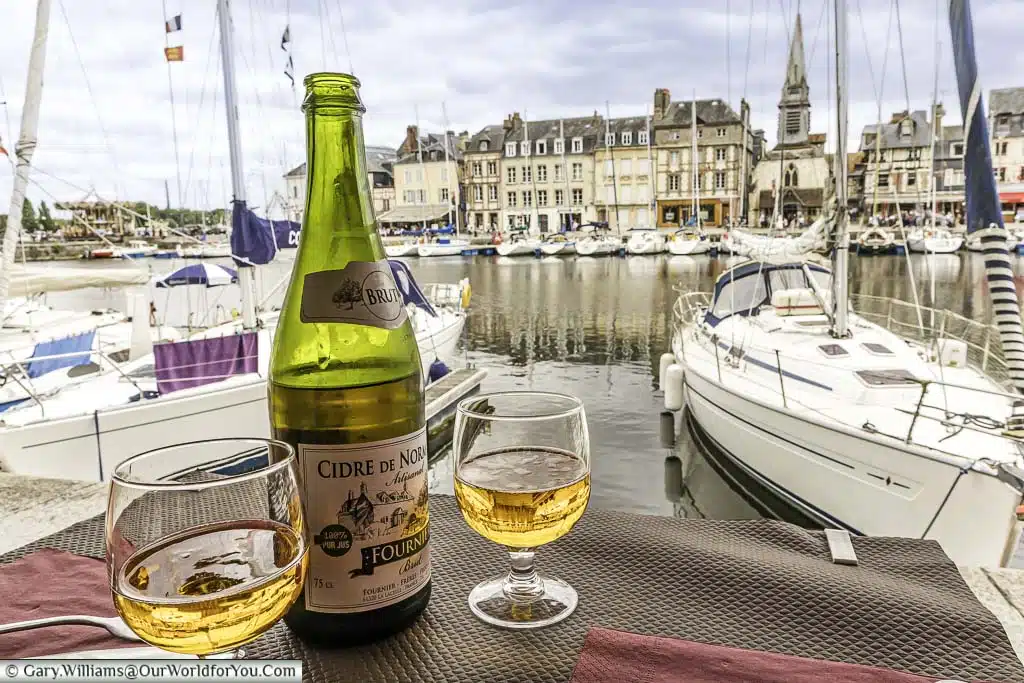 A bottle and 2 glasses of Cidre de Normandie served at the edge of the marina of Honfleur, filled with small sailing boats.