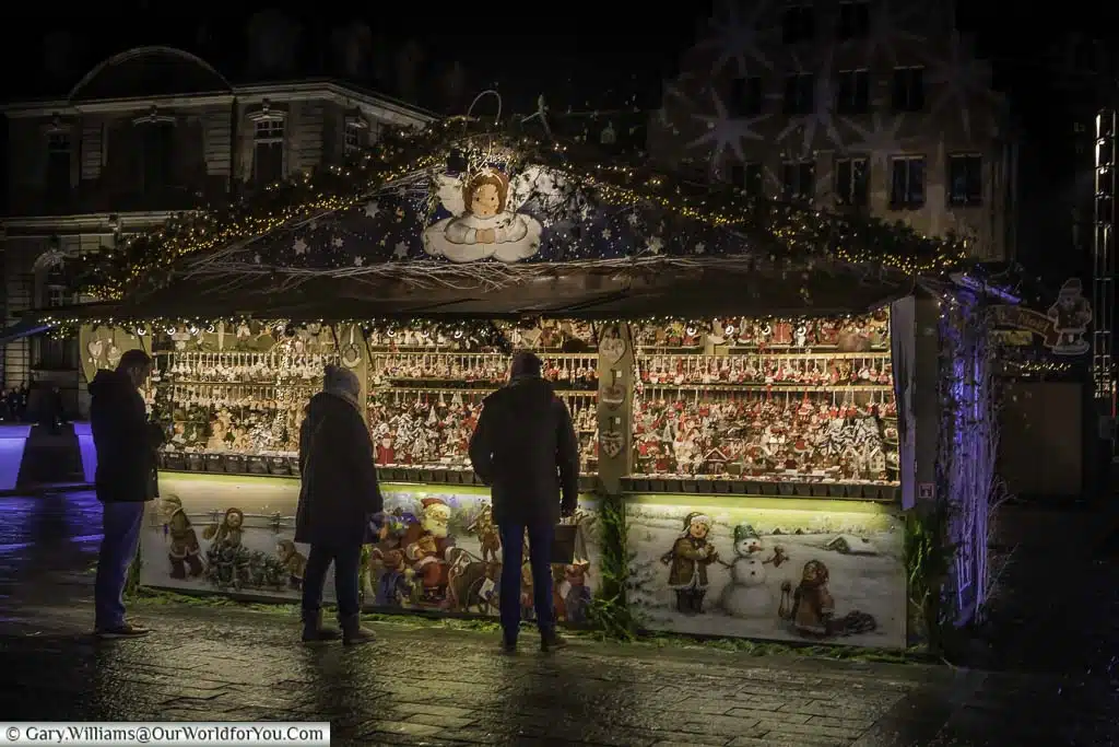 A Christmas market stall in the Place De La Cathédrale decorated with Christmas scenes of Santa and his sleigh, children with a snowman and an angel looking down at the shoppers perusing its extensive range of Christmas decorations.