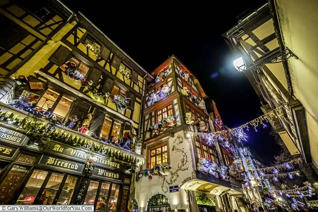 Looking up along Rue du Maroquin at night full of traditional half-timbered buildings that look extra special at Christmas. Lights hang between the buildings and every window decorated with lights and one building, teddy bears.