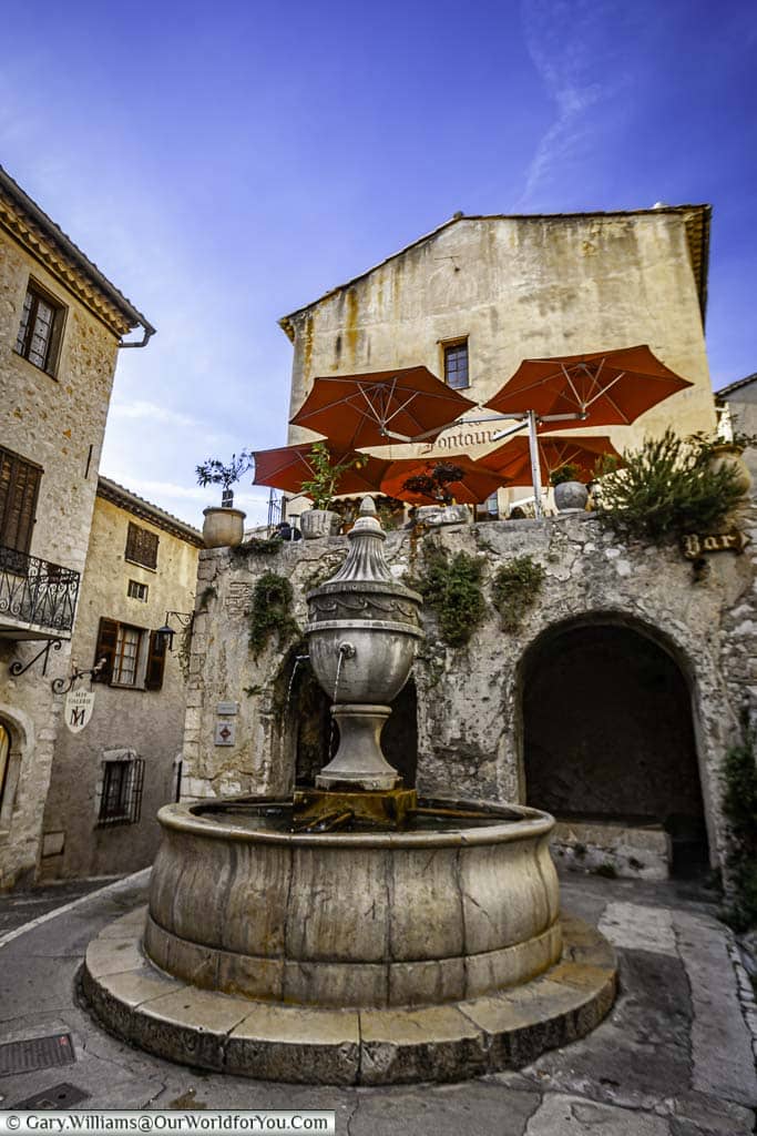 A stone fountain in a courtyard of the provencal village of st paul de vence, below the red parasols of a roof top cafe under deep blue skies