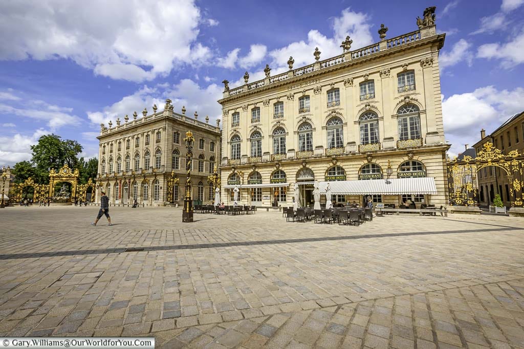 A view from the centre of the square Place Stanislas with three storey buildings with cafes at the bottom. You can also see the black & gold wrought iron gates and lamp posts.