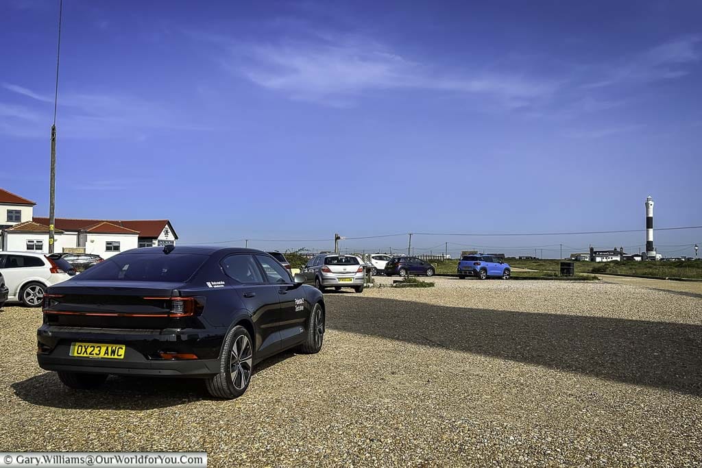 Our black Polestar 2 in a gravel car park with Dungeness Lighthouse in the distance