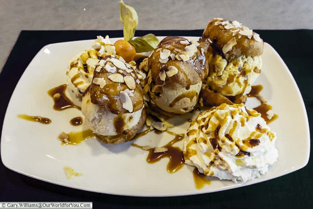 Three profiteroles stuffed with different flavoured ice creams topped with toasted almonds with a dollop of whipped cream on the side, all drizzled with caramel sauce.
