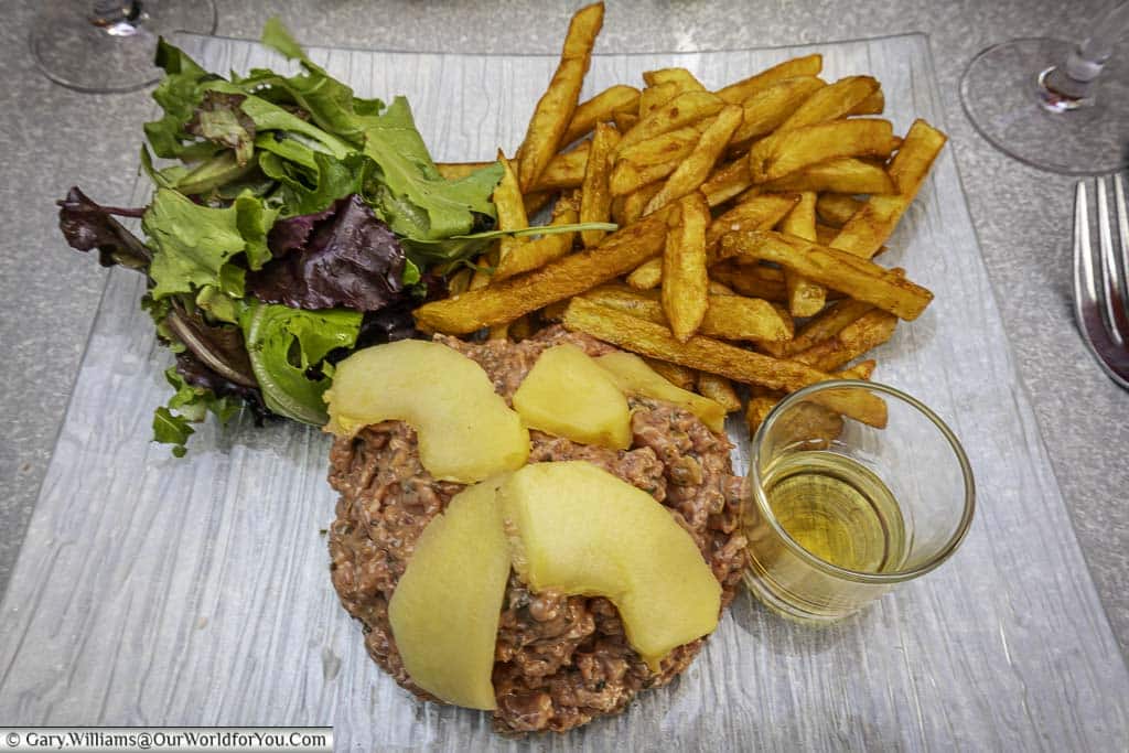 A plate of steak tartare served with a side salad and fried potatoes. this dish has a regional twist in that is covered with poached apples and shot of calvados spirit on the side