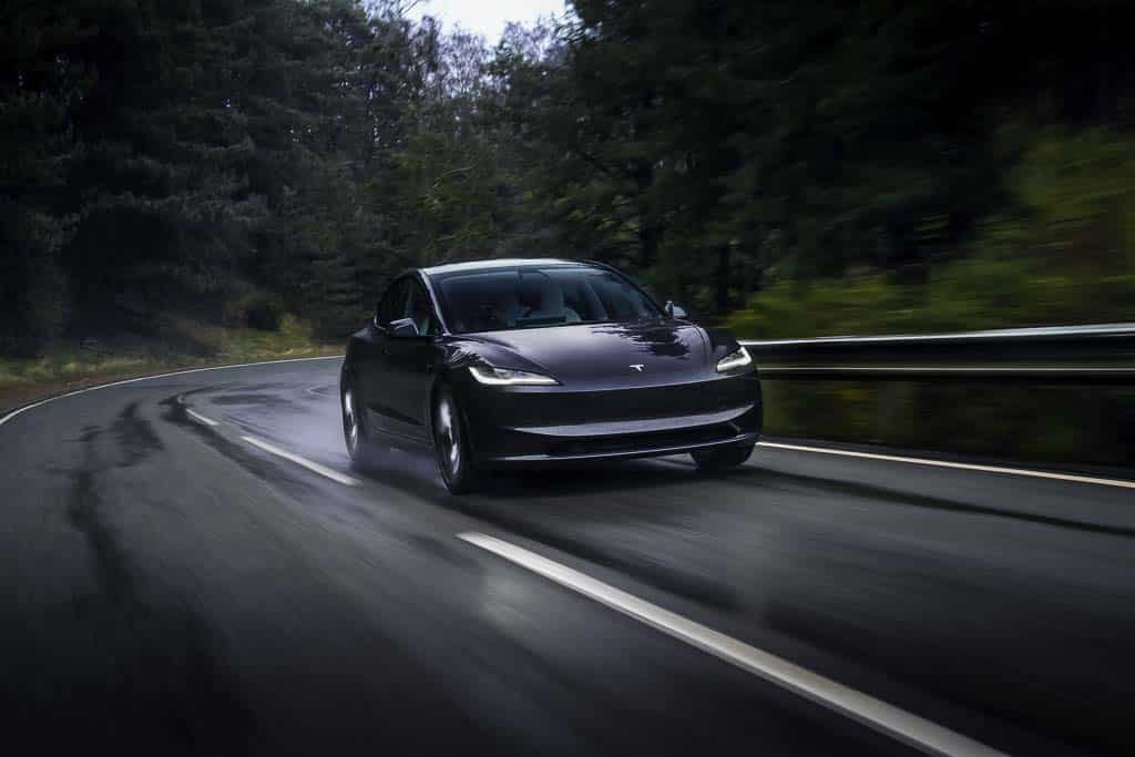 A stock photo ofa tesla model 3 driving along a damp road thtrough the forest