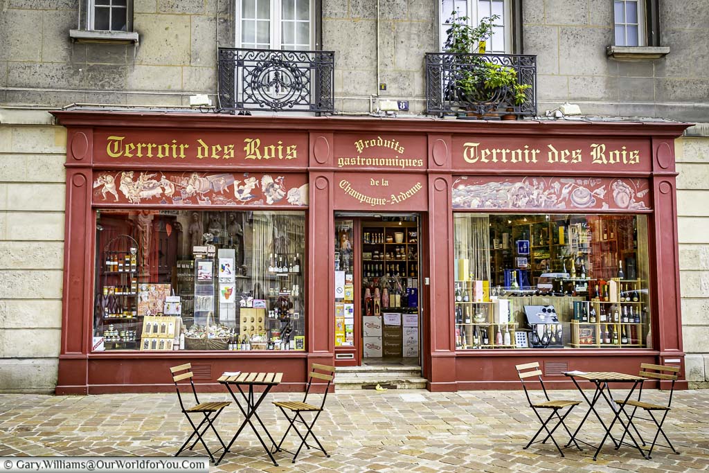 2 tables and chairs outside a gift shop painted in deep red, providing local gastronomic food produce from the Champagne-Ardenne region. The shop is called Terroir des Rois which roughly translates I from the soil of Kings.