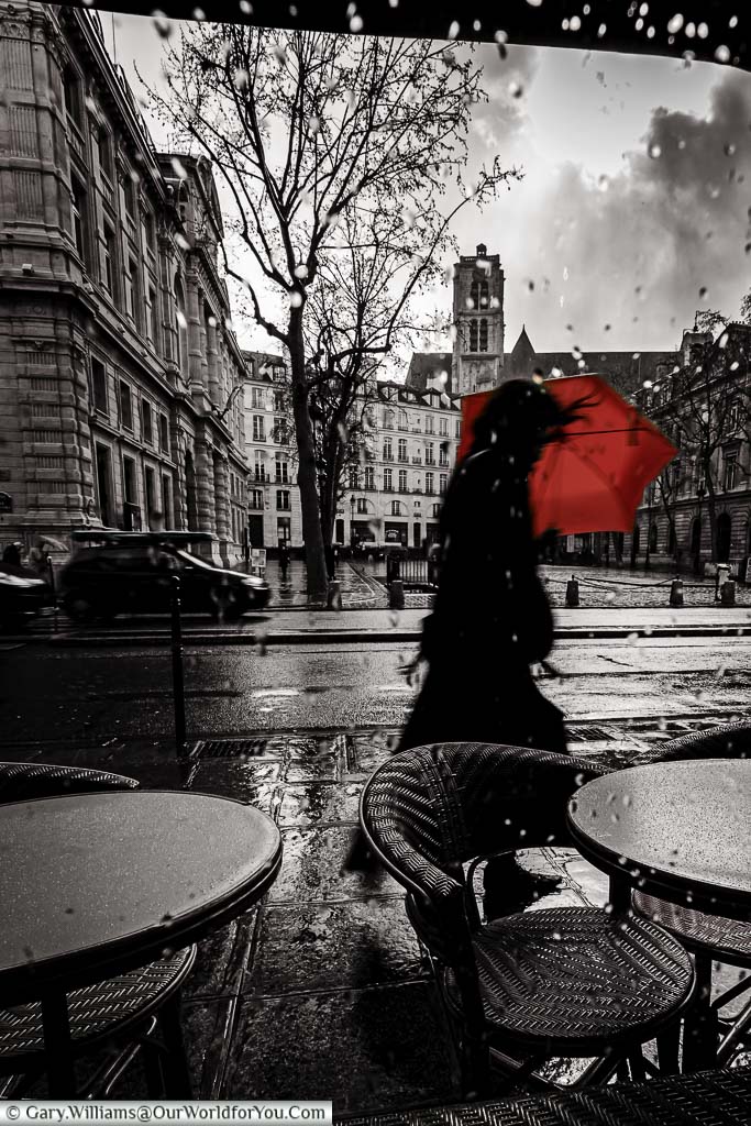 A black and white image, taken from inside a café, of a woman passing by with red umbrella, wrestling against the elements, on a stormy day in Paris