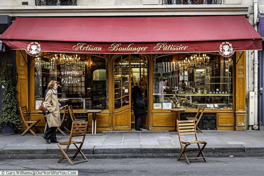 An elegant woman passing by I traditional artisan boulanger pâtissier on side road in Paris. The front of the art deco styled shop is panelled in wood with 2 large windows displaying chandeliers inside, there is a deep burgundy canopy covering the upper portion of the store.