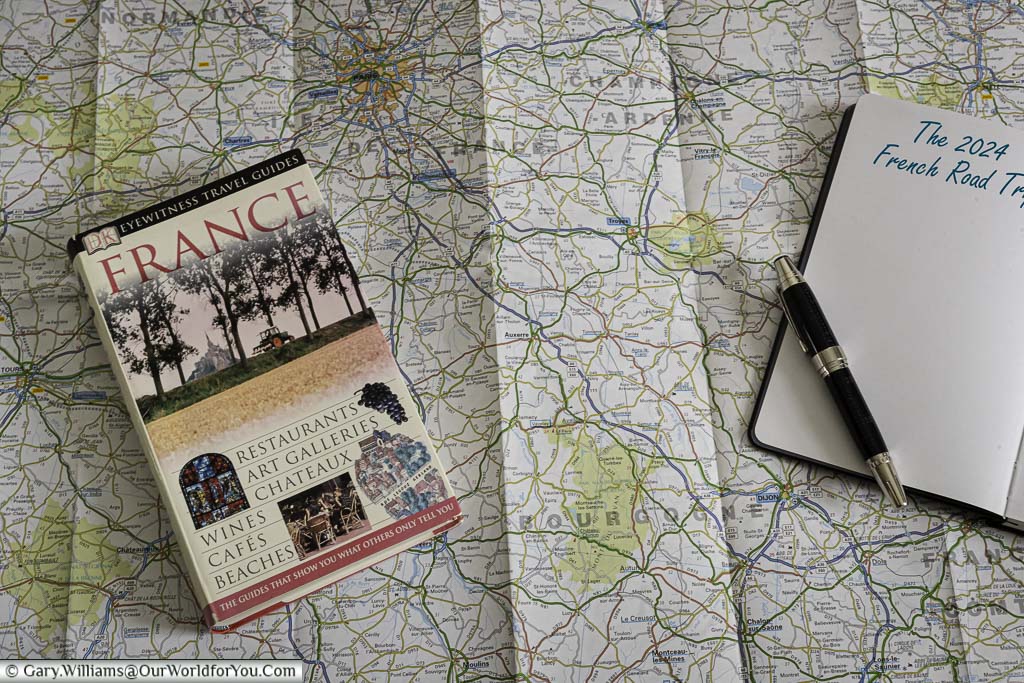 A french guide book, a fountain pen resting on a notepad with the page titled 'the 2024 french road trip' on top of a folded out aa road map of france