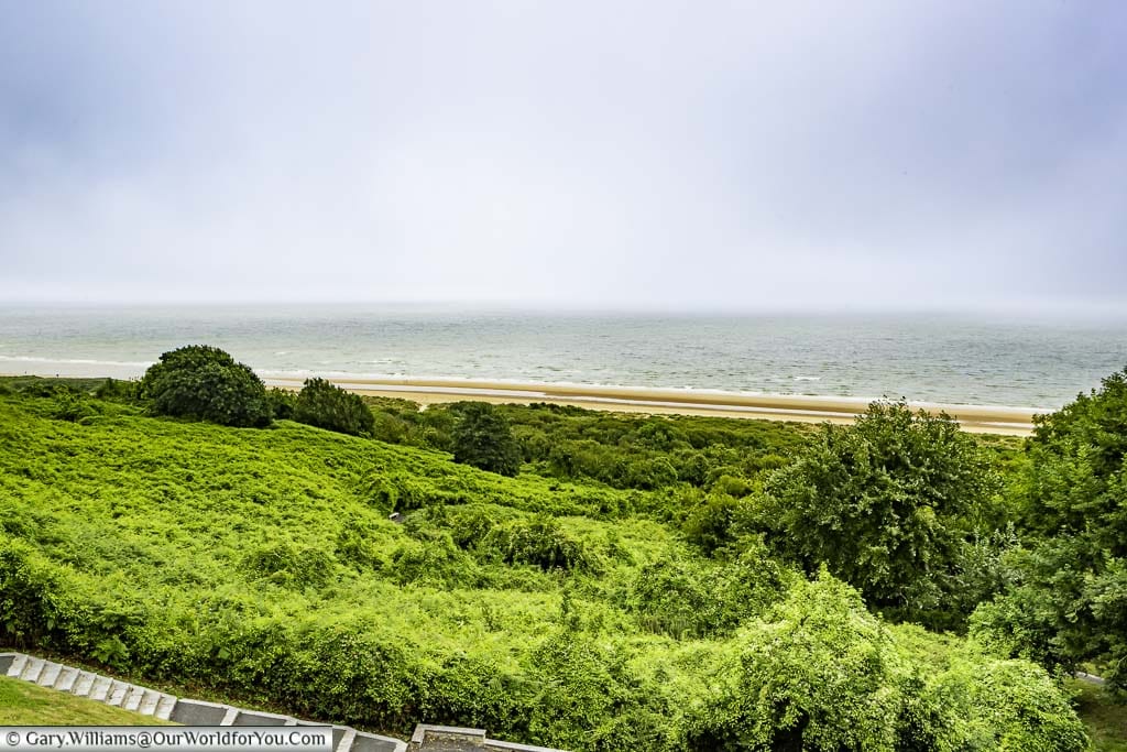 A view of the shoreline of 'Omaha' beach in Normandy, featuring a grassy mound, a thin strip of sand, and the ocean beyond.