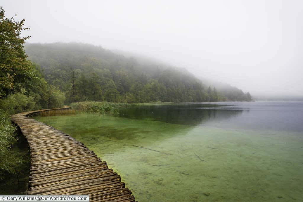 A damp path, constructed of wooden planks, skirts the end of one of Plitvice's many lakes, framed by a tree-covered hill. A mist hangs over the lake and clings to the hillside.
