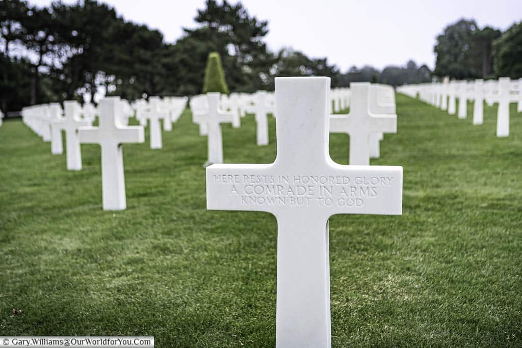 A simple white marble cross dedicated to an unknown soldier in front of rows of further headstones in the Normandy American Cemetery