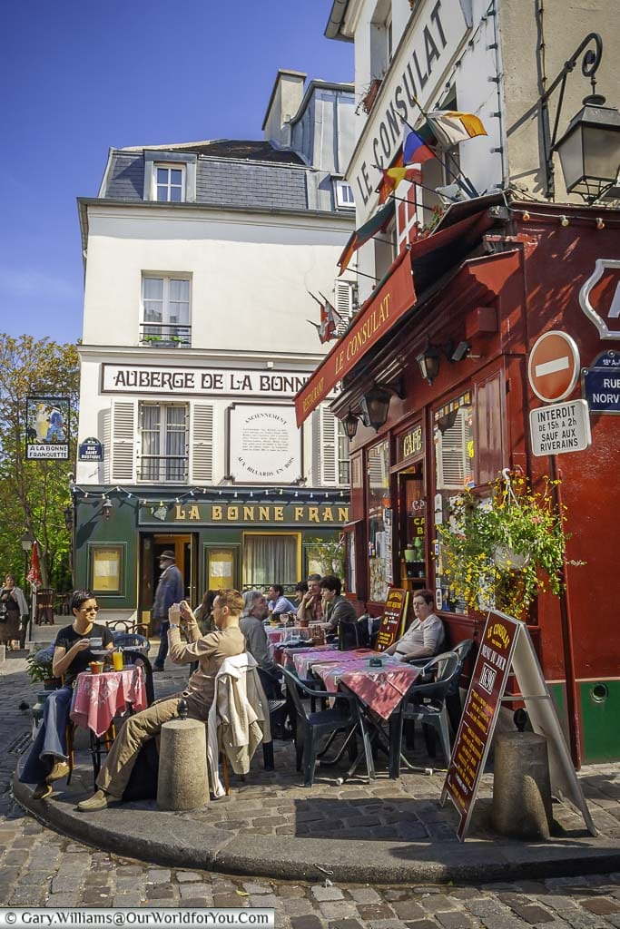 A street scene in Montmartre outside Le Consulat on a bright sunny day with people enjoying lunch at tables outside the restaurant.