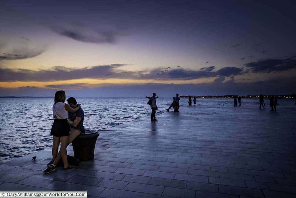 Individuals and couples on the zadar’s riva at dusk, listening to the sea organs as the adriatic sea laps against the edge.