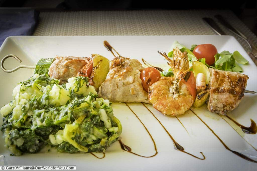 A fish skewer served with potatoes & greens, a light salad on a white rectangular plate.