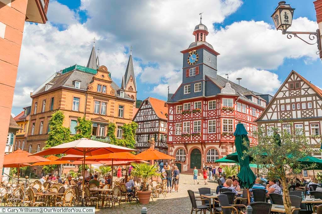 The bustling marktplatz, the centre of the Old Town of Heppenheim. Around the edges of the square tables and chairs under parasols for the local cafes and bars. In the background is the town Hall another half-timbered building with a tiled clocktower.