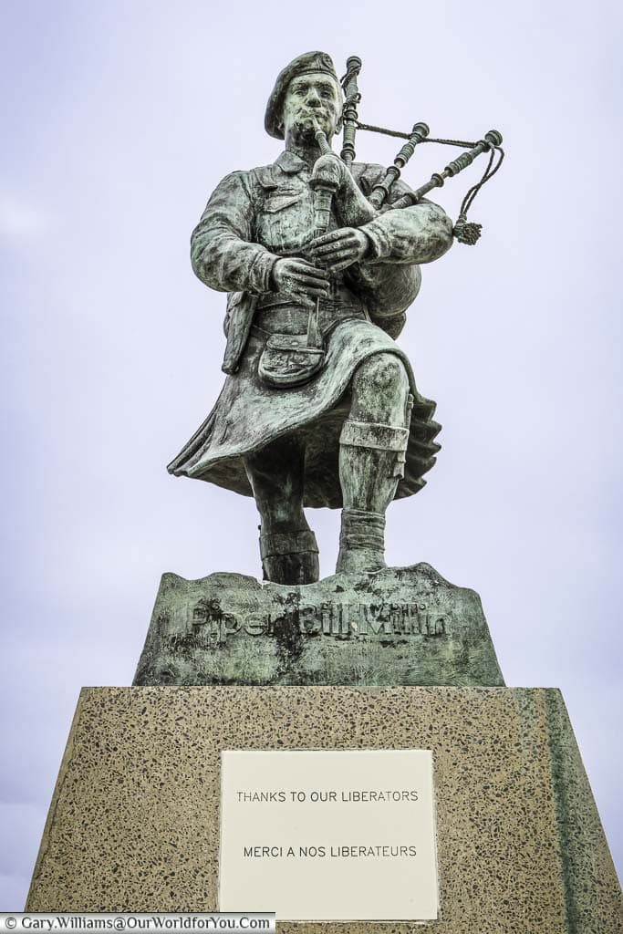 A brass statue of the scottish soldier, and regimental piper, bill millin, playing bagpipes as he strides up sword beach in normandy. The plinth has a plaque at that reads 'thanks to our liberators' in both english and french
