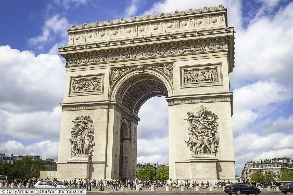 A close-up shot of the Arc de Triomphe in the centre of Paris's infamous roadabout taken on a bright day with bright white clouds scattered across a blue sky.