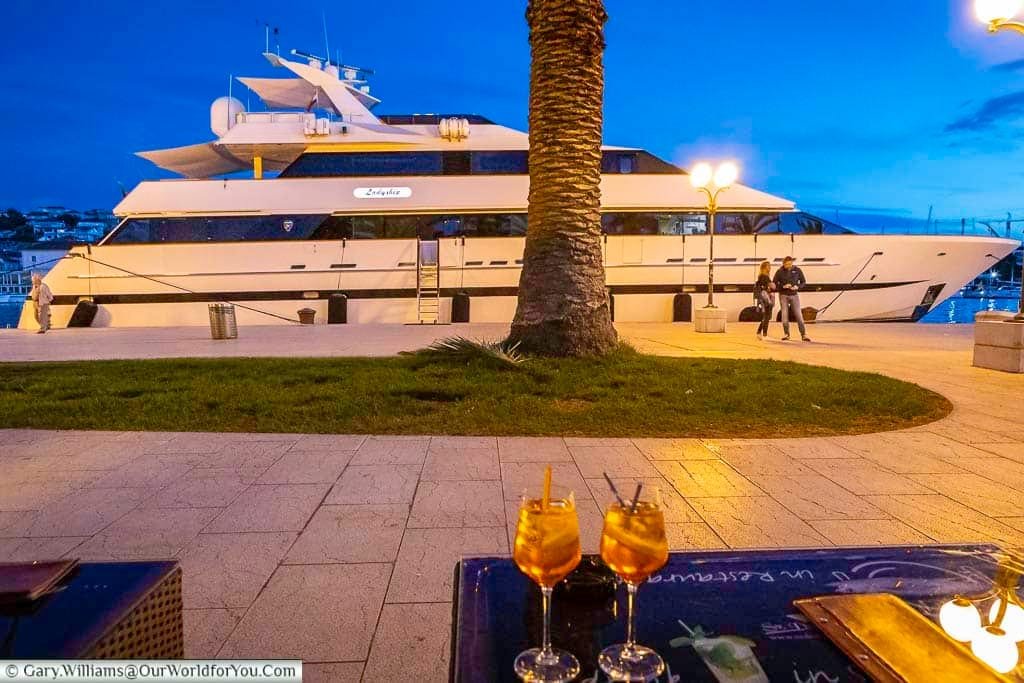 two glasses of aperol spritz at a bar on the riva, with a lager motor cruiser moored in the background of trogir, croatia