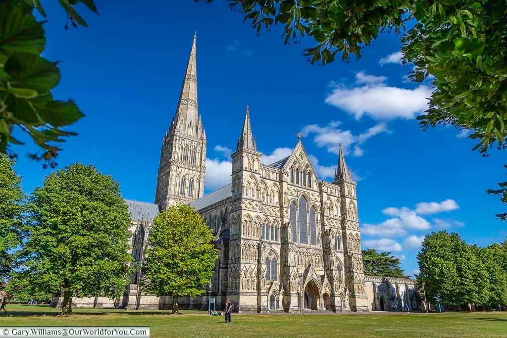 The view of the west end of salisbury cathedral, showing its impressive spire and gothic architecture glistening in the sunlight under a blue sky