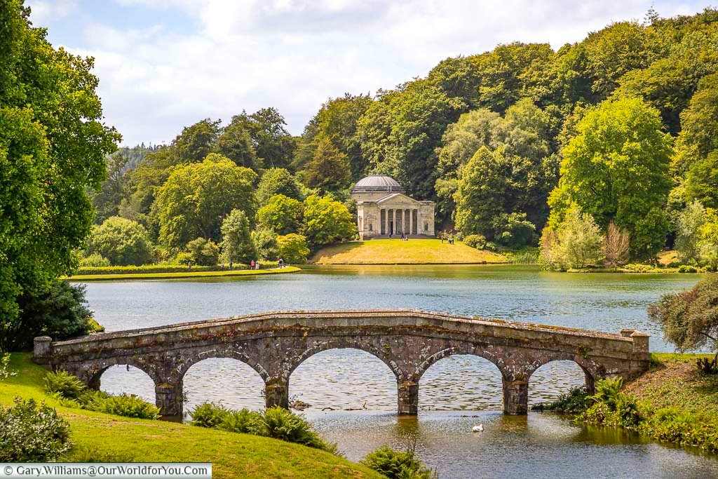 The palladian bridge and pantheon at stourhead next to the edge of the lake, set in lush green woodland on a bright day