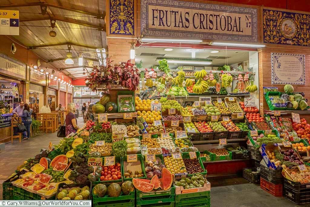 Inside the Mercado de Triana. A vendors booth, decorated with traditional tiles of the region, is piled high with fruit and vegetables.