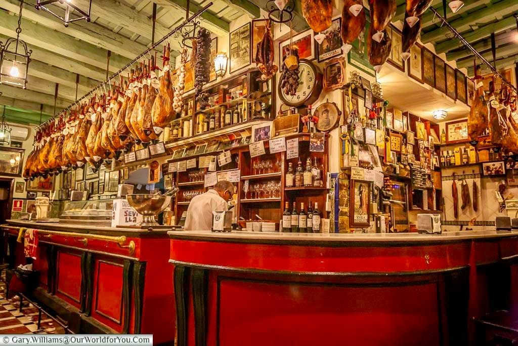 A mature bartender stands behind a traditional marble-topped counter in a bar in the Barrio Santa Cruz area of Seville. Legs of Iberico ham hang from the ceiling rafters.