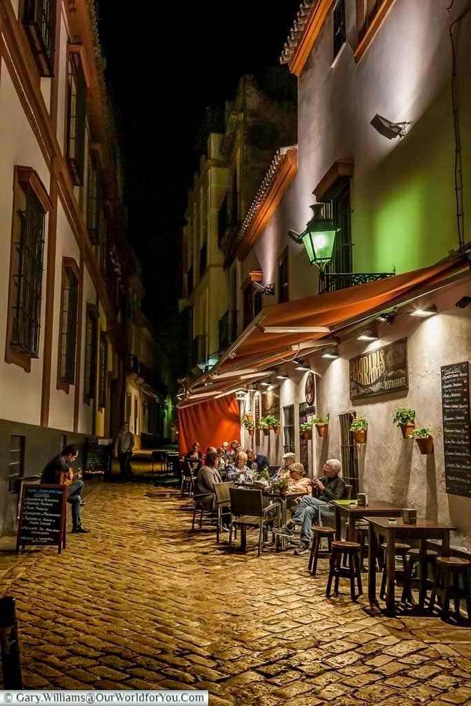 A musician plays a Spanish guitar, in a narrow cobbled lane, in front of a group of people sitting outside a restaurant at night in the Barrio Santa Cruz area of Seville.