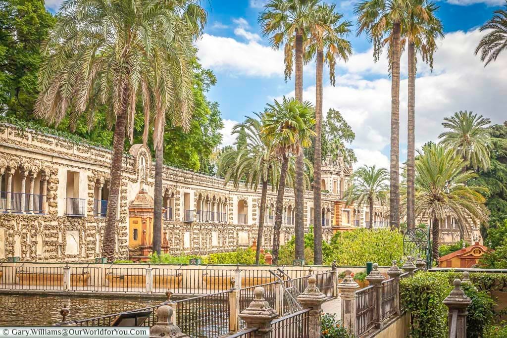 Tall palm trees dominate the view with an ornate high wall as a boundary to the gardens of the Alcazar of Seville.