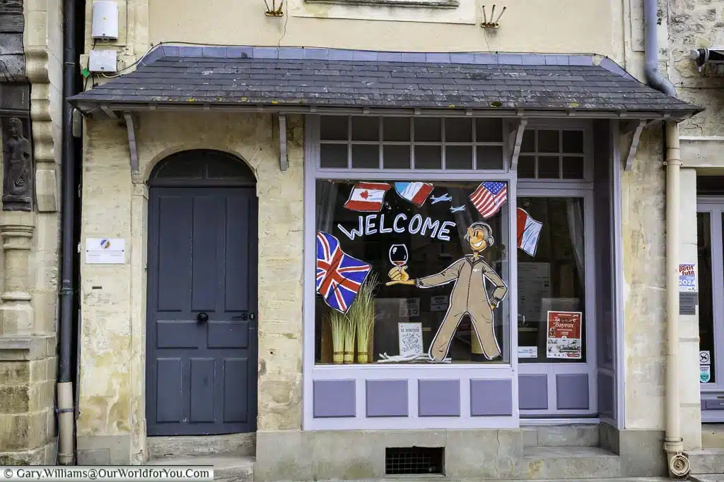 a quaint shop front where the windows are decorated with painted flags of the allied powers and an airman holding a glass of red wine with the words welcome in bayeux, normandy