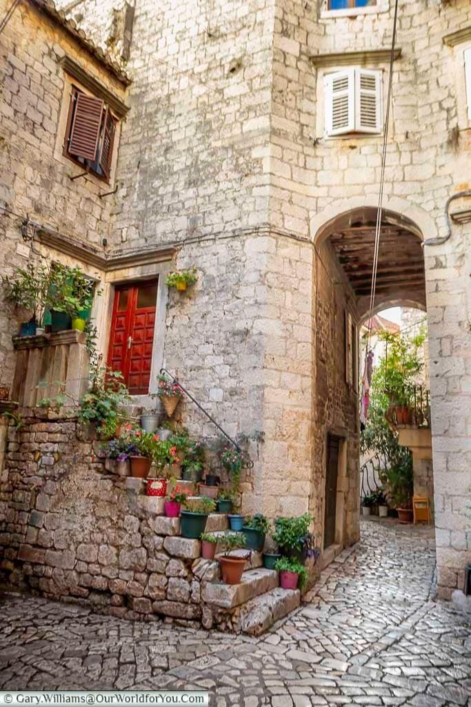 A small courtyard with cobbled paths, stop steps leading to the buildings made from golden-coloured stone. Flowerpots decorate the steps and provide the colour in the scene.