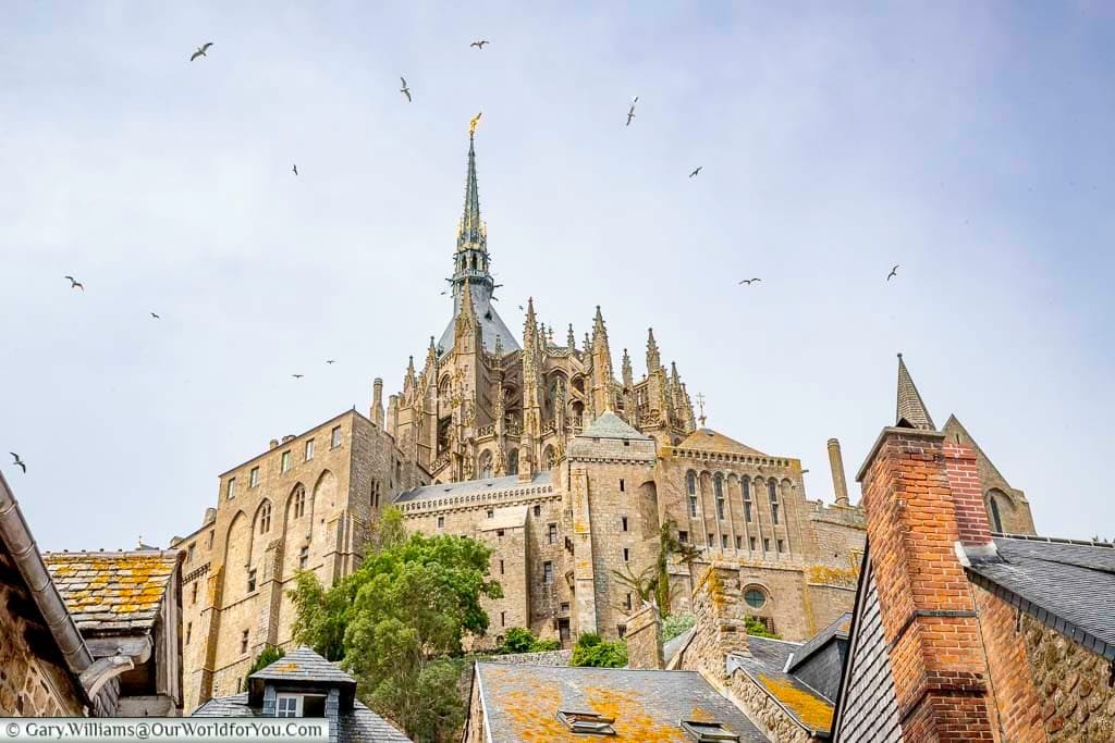 Birds circle the spire of the Mont-Saint-Michel Abbey, high on the hill, as the skies darken.