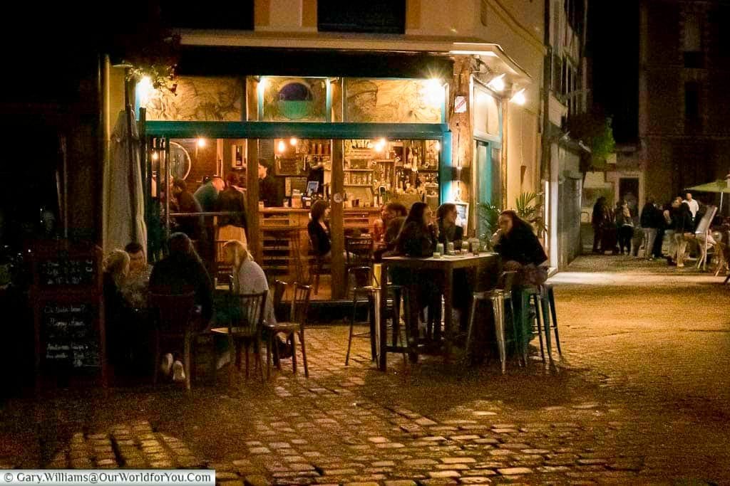 An evening scene of people sitting both inside and outside a cafe/bar in Rouen.