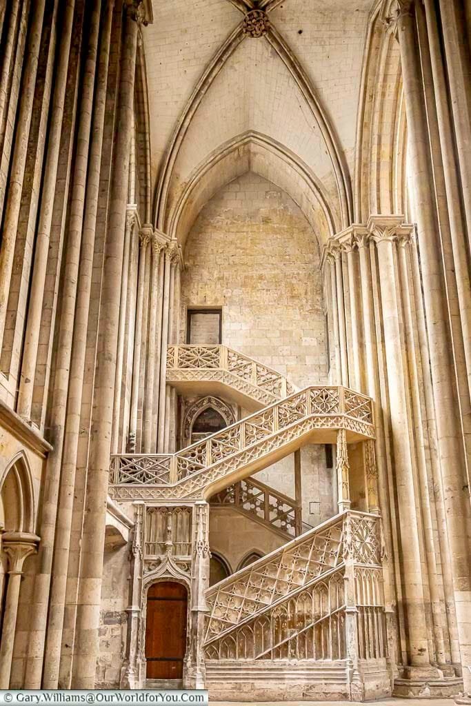 A portrait view of an elegant stone staircase, within Rouen Cathedral in Normandy, leading from a wooden door at ground level up two floors. This ornate piece is known as the bookseller's staircase.