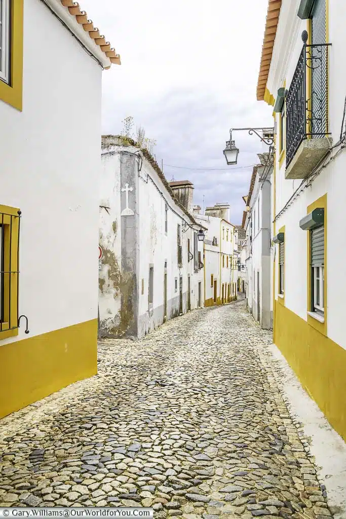A narrow cobbled lane between white buildings, trimmed with a bold yellow, in the city of Évora, Portugal