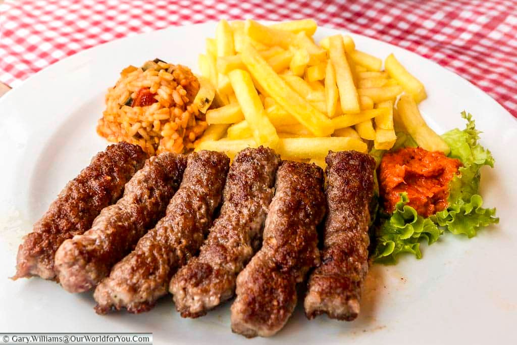 A white plate showcases a generous serving of six grilled cevapcici sausages, accompanied by a pile of golden French fries, a small mound of red pepper relish (ajvar), a dollop of vibrant red rice with vegetables, and a few fresh lettuce leaves. The meal is presented on a table covered with a red and white checkered tablecloth, suggesting a casual dining setting.