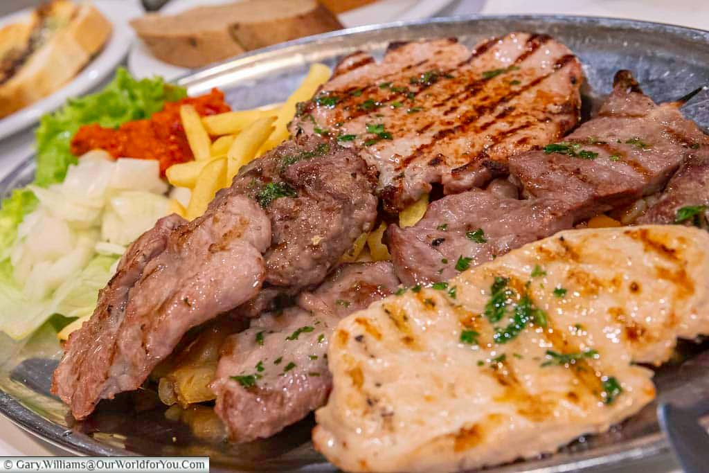 A silver platter is filled with a variety of grilled meats, including pork chops and other cuts, generously seasoned with herbs and displaying distinct grill marks. The platter also includes golden French fries, chopped white onions, a dollop of vibrant red ajvar relish, and fresh green lettuce, suggesting a hearty and flavorful Croatian mixed grill.
